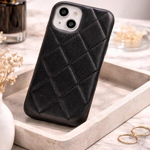 May include: Black quilted leather phone case with camera cutouts. The case is on a white tray with gold rings and a perfume bottle in the background. The OpenWillow logo is visible in the top right corner.