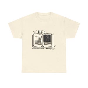 Puede incluir: Camiseta color crema con un gráfico negro de un libro abierto. El texto "U.C.K." está arriba y "Unlimited Cosmic Knowledge" abajo. El diseño incluye estrellas y una ilustración detallada dentro del libro.