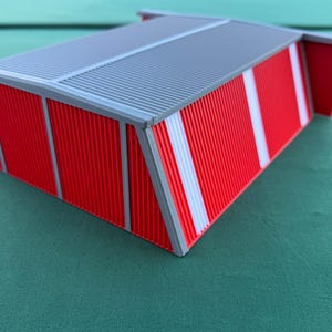 May include: A miniature red and gray model of a building with a corrugated metal roof. The building has red walls with white vertical stripes and gray trim. The model is likely for architectural or hobbyist use.