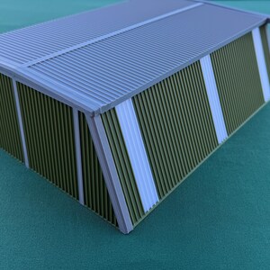 May include: A miniature model of a building with a corrugated metal roof and walls. The walls are olive green with vertical white stripes. The roof and trim are gray.