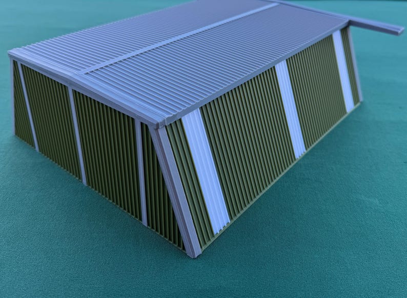 May include: A miniature model of a building with a corrugated metal roof and walls. The walls are olive green with vertical white stripes. The roof and trim are gray.