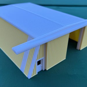 May include: A miniature model of a yellow and white building with a corrugated roof. The building has a rectangular shape with a white door and a white overhang. The walls are yellow with white vertical stripes.