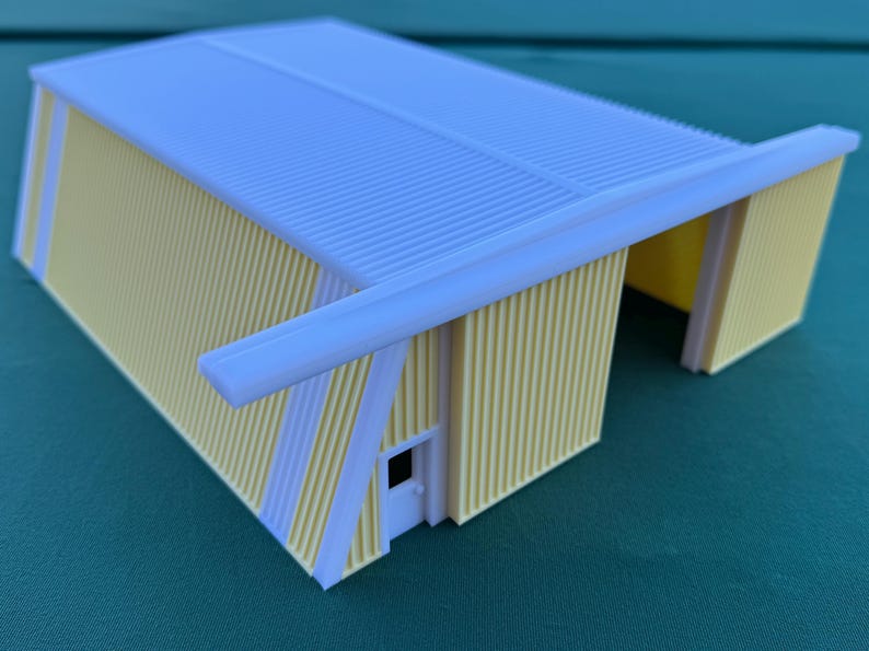 May include: A miniature model of a yellow and white building with a corrugated roof. The building has a rectangular shape with a white door and a white overhang. The walls are yellow with white vertical stripes.