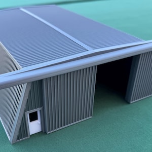 May include: A gray model of a corrugated metal building with a white door. The building has a sloped roof and an open front. The model is on a green surface.