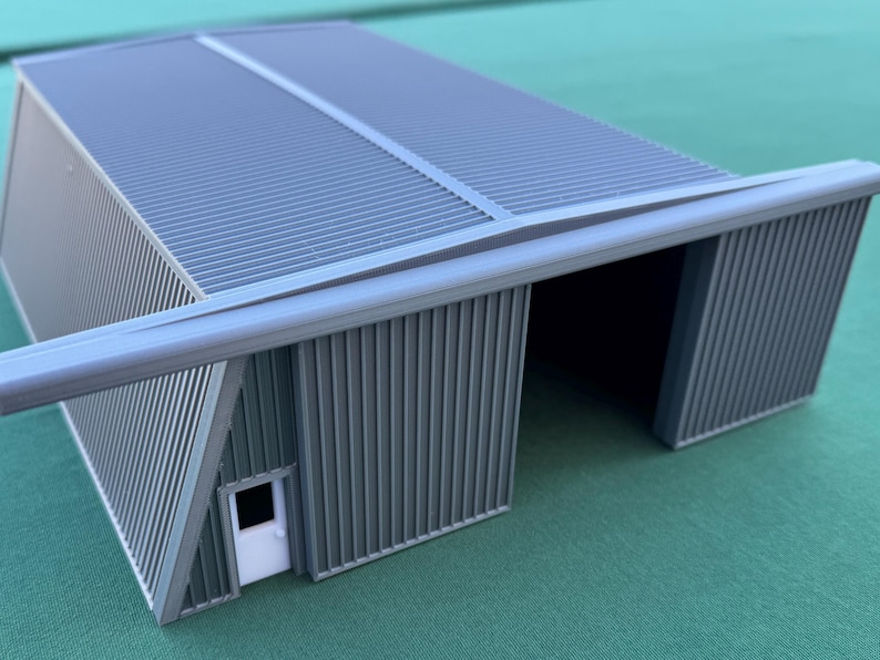 May include: A gray model of a corrugated metal building with a white door. The building has a sloped roof and an open front. The model is on a green surface.