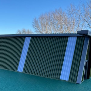 May include: A miniature green and blue corrugated metal shed model. The shed has vertical ridges and two vertical blue stripes. The roof and trim are dark gray. The model is on a teal surface.