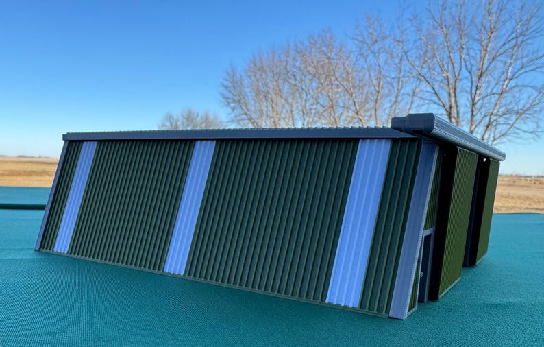 May include: A miniature green and blue corrugated metal shed model. The shed has vertical ridges and two vertical blue stripes. The roof and trim are dark gray. The model is on a teal surface.