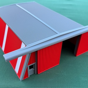 May include: A red and gray model of a hangar or industrial building. The building has a corrugated metal roof and walls with white stripes. A small door is visible on one side.