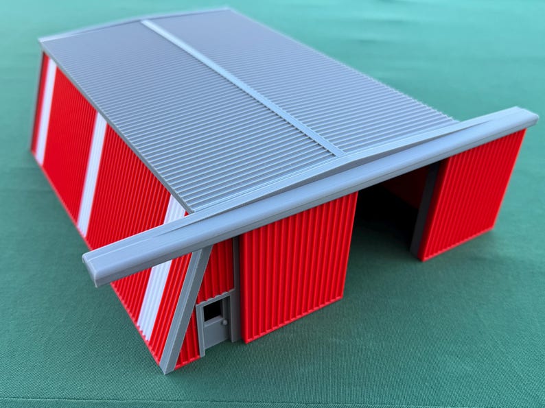 May include: A red and gray model of a hangar or industrial building. The building has a corrugated metal roof and walls with white stripes. A small door is visible on one side.