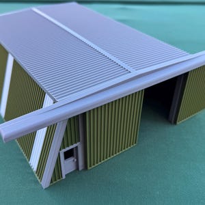 May include: A miniature model of a corrugated metal building with a gray roof and olive green walls. The building has a small door and white accent stripes. The model is a detailed representation of a hangar or warehouse.