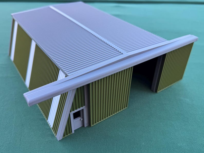 May include: A miniature model of a corrugated metal building with a gray roof and olive green walls. The building has a small door and white accent stripes. The model is a detailed representation of a hangar or warehouse.