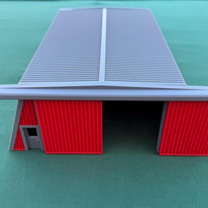 May include: A model of a gray hangar with a corrugated roof and red doors. The building has a rectangular shape with a small door on the side. The hangar is set against a green background.