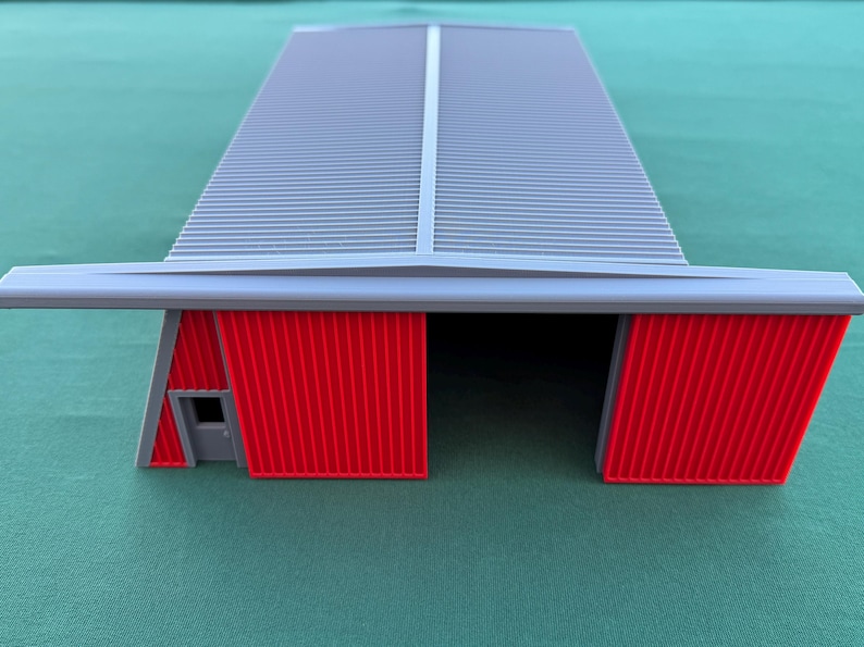 May include: A model of a gray hangar with a corrugated roof and red doors. The building has a rectangular shape with a small door on the side. The hangar is set against a green background.