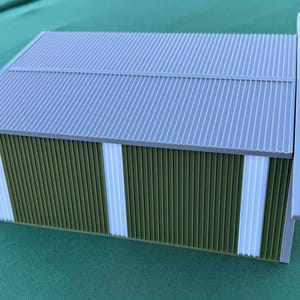 May include: A miniature model of a building with a corrugated gray roof and green and white walls. The building has a rectangular shape and a white trim along the edges. The model is set against a green background.