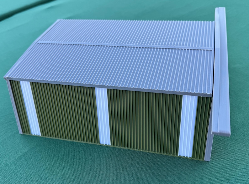 May include: A miniature model of a building with a corrugated gray roof and green and white walls. The building has a rectangular shape and a white trim along the edges. The model is set against a green background.