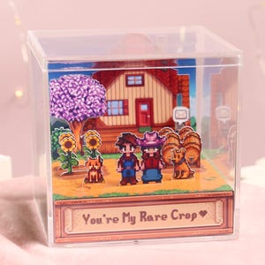 Puede incluir: Caja de acr&iacute;lico transparente con un diorama pixelado. La escena incluye una casa, girasoles, un &aacute;rbol y dos figuras tomadas de la mano. El texto "You're My Rare Crop" se muestra en una pancarta en la parte inferior.