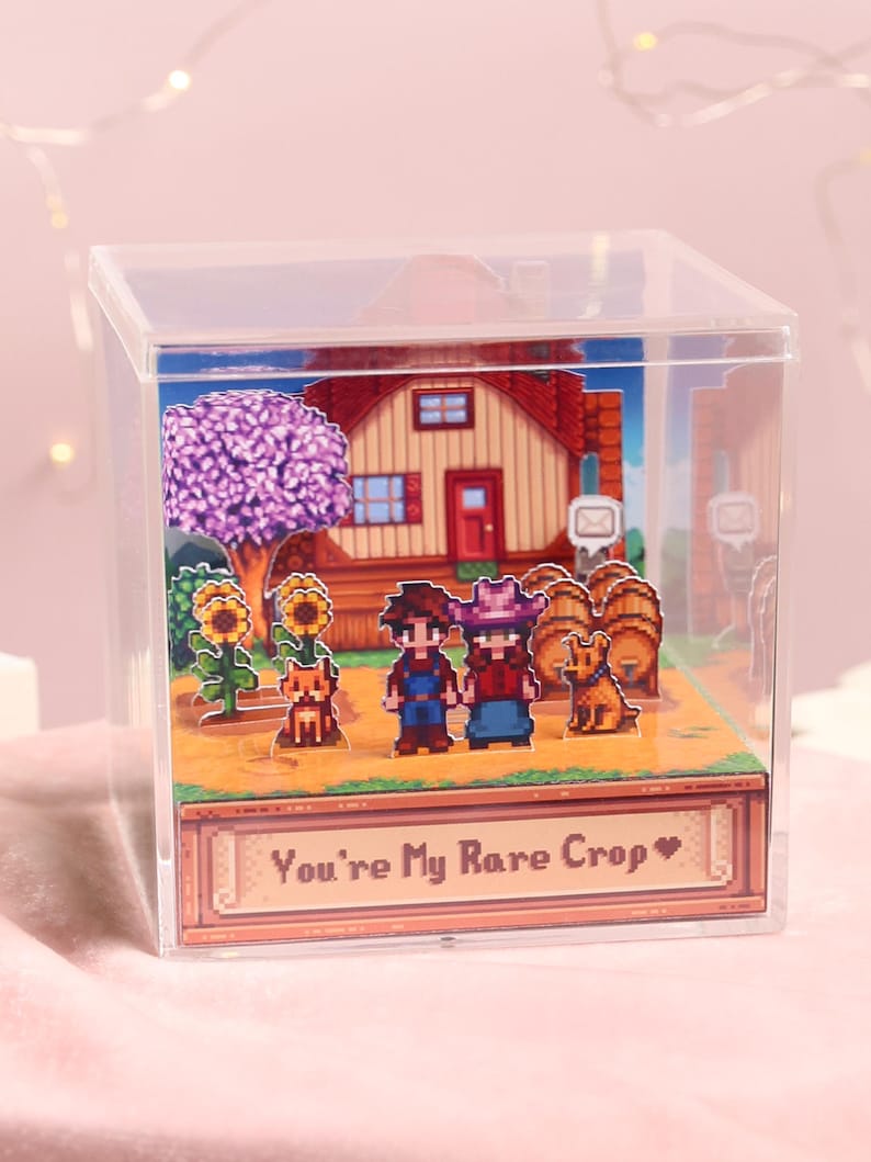 Puede incluir: Caja de acr&iacute;lico transparente con un diorama pixelado. La escena incluye una casa, girasoles, un &aacute;rbol y dos figuras tomadas de la mano. El texto "You're My Rare Crop" se muestra en una pancarta en la parte inferior.