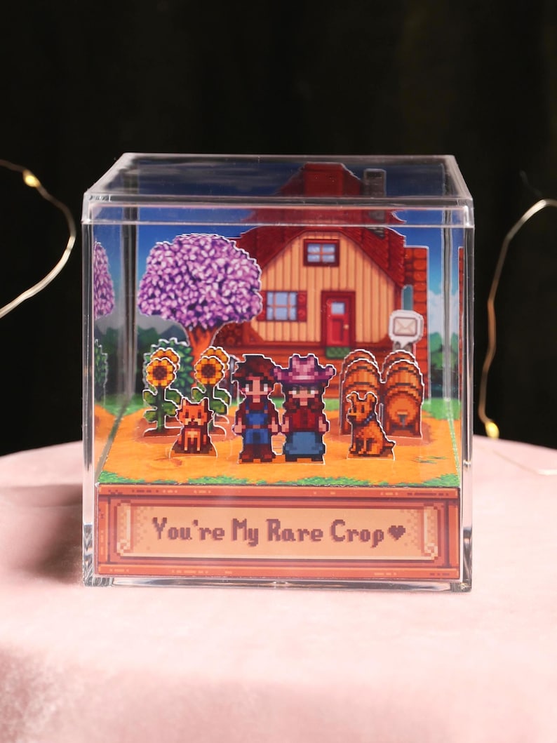 Puede incluir: Un diorama c&uacute;bico de acr&iacute;lico transparente con una escena de granja pixelada. La escena incluye una casa, &aacute;rboles, girasoles, dos personajes, dos perros y barriles. La base del cubo tiene el texto "You're My Rare Crop" en una pancarta decorativa.