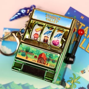 Könnte beinhalten: Ein Miniatur-Schlüsselanhänger in Form eines Spielautomaten in Metallic-Grün mit dem Schriftzug "Stardew Valley" oben. Die Walzen zeigen spielbezogene Symbole. Der Boden der Maschine zeigt eine Landschaft. Ein roter Griff und ein blauer Anhänger sind ebenfalls sichtbar.