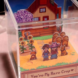 Puede incluir: Una caja de acr&iacute;lico transparente con un diorama en miniatura de una escena de granja pixelada. La escena incluye una casa con techo rojo, girasoles, personajes y el texto "You're My Rare Crop".