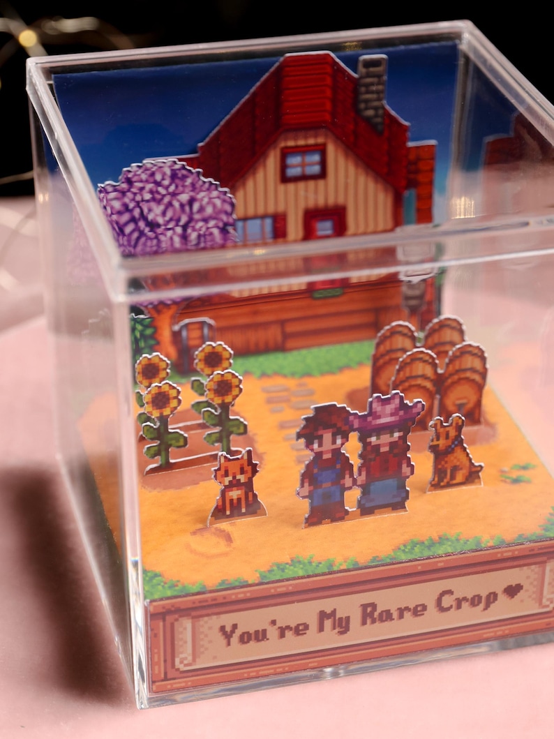 Puede incluir: Una caja de acr&iacute;lico transparente con un diorama en miniatura de una escena de granja pixelada. La escena incluye una casa con techo rojo, girasoles, personajes y el texto "You're My Rare Crop".
