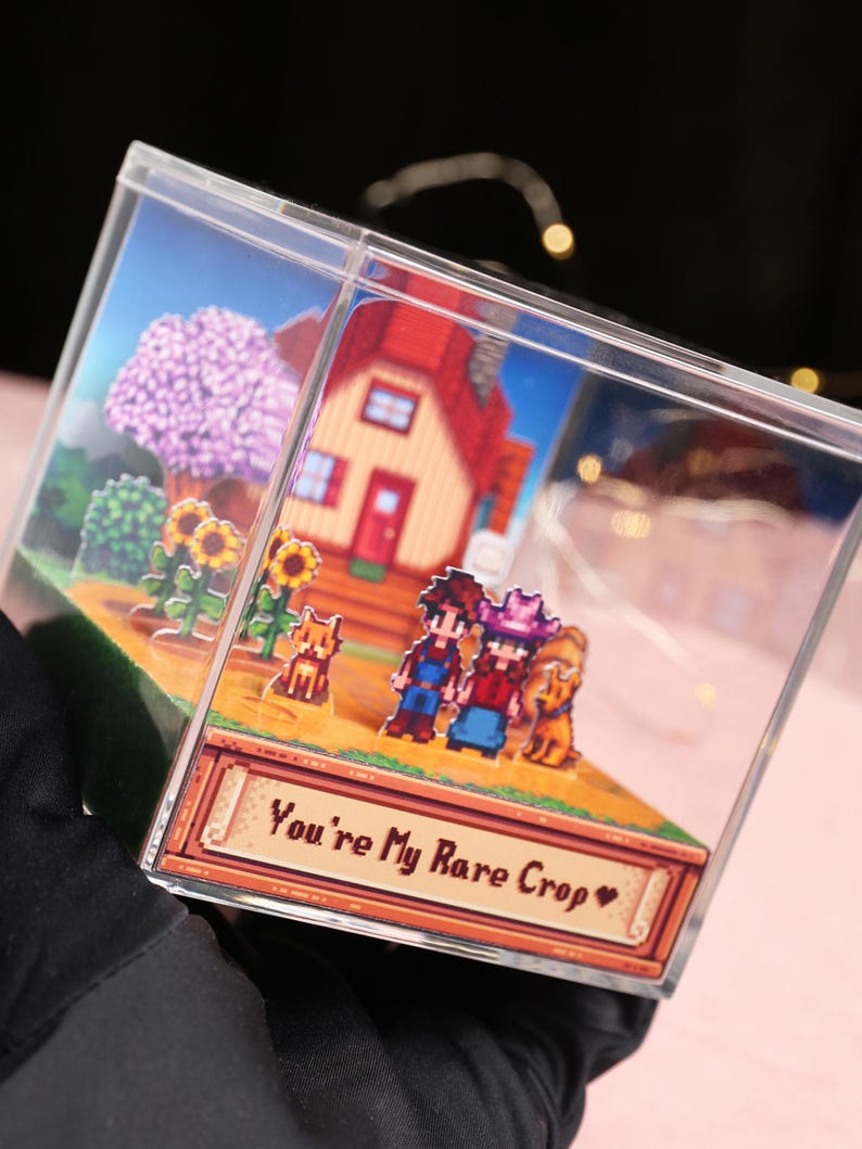 Puede incluir: Una caja de acr&iacute;lico transparente que presenta una escena de diorama pixelada con una casa de techo rojo, personajes y el texto "You're My Rare Crop". La escena incluye girasoles, &aacute;rboles y un perro. La caja es sostenida por una mano enguantada.