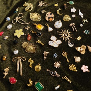 Può includere: Una collezione di vari ciondoli e pendenti dorati e colorati, tra cui cuori, fiori, animali e forme geometriche. I ciondoli sono sparsi su una superficie di velluto verde scuro, mettendo in risalto i loro intricati disegni e dettagli.