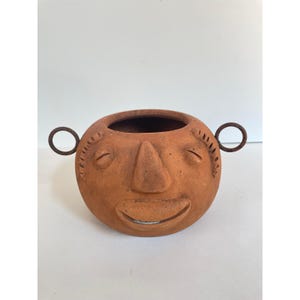 Puede incluir: Una maceta redonda de cerámica de color terracota con un diseño de cara sonriente. La maceta tiene una nariz, ojos cerrados y una boca ancha y abierta. Dos asas de anillo de metal están unidas a los lados. Adecuada para plantas pequeñas o decoración.