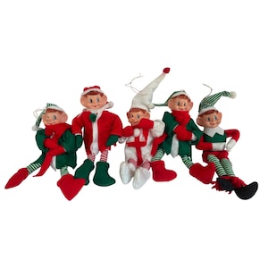 May include: Five elf dolls with painted faces and pointed ears. The dolls wear fabric outfits in red, green, and white, featuring striped leggings, pointed hats, and scarves. Each doll has a small string loop attached to the top of its hat for hanging.