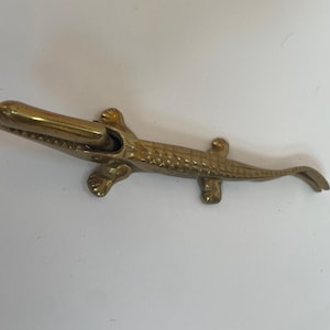 May include: A brass alligator-shaped bottle opener. The opener is detailed with textured scales and features a hinged jaw. The alligator is lying down on a white surface. The opener is made of a shiny, gold-colored metal.