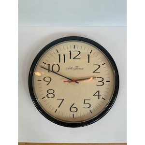 May include: A round Seth Thomas wall clock with a black frame and a cream-colored face. The clock features black numerals, a black minute hand, a black hour hand, and a red second hand. The clock is approximately 12 inches in diameter.