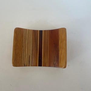 May include: A wooden coaster with a curved design. The coaster is made from various wood types, featuring a mix of light and dark brown tones, with vertical stripes and solid sections. The edges are rounded.