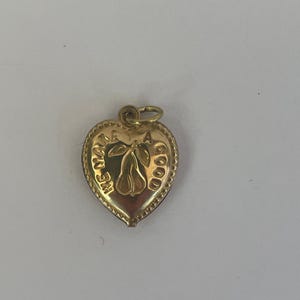 May include: A gold-colored heart-shaped pendant with a pear illustration in the center. The pendant has the words "WE MAKE A GOOD" around the pear. The pendant has a small loop at the top for a chain.