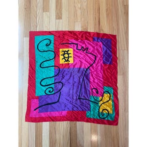 May include: A vibrant silk scarf with an abstract design. The scarf features a red border framing a central composition of pink, purple, teal, and yellow geometric shapes. Black lines and shapes add further detail to the design. The scarf is a square shape.
