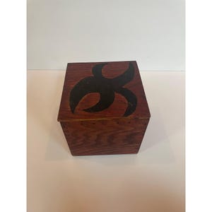 May include: A wooden box with a dark brown finish. The top of the box features a black abstract design. The box is a cube shape and appears to be handmade.