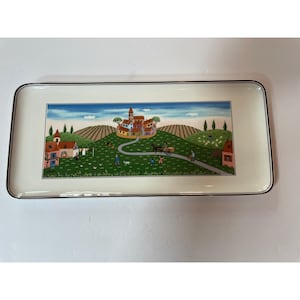 May include: Rectangular ceramic serving tray with a cream-colored background and a painted village scene. The scene depicts a village with red-roofed buildings, rolling green hills, and a winding road. The tray has a black border.