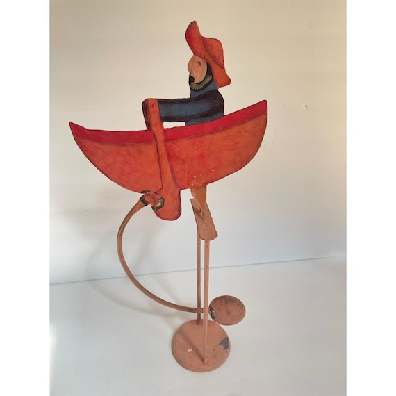 A hand-painted vintage folk-art fisherman in a rowboat that balances and rocks via counterweight — the kind of tactile, characterful object you'd find at a quirky antique shop and never forget.