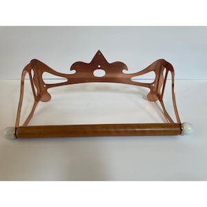 May include: A copper-colored metal paper towel holder with a decorative top and a wooden dowel. The holder has white ceramic ball finials on each end of the dowel. The design is vintage and elegant.
