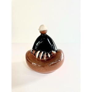 May include: A ceramic figurine featuring a rounded brown base, a glossy black upper section, and a small white cap. The figure displays a seated posture with arms crossed, accented by white and black stripes on the sleeves, creating a smooth, reflective surface finish throughout the entire object.