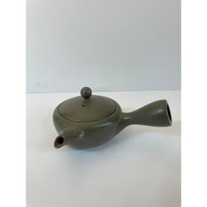 May include: Olive green ceramic teapot with a side handle and a small, round lid. The spout is small and angled. The teapot is on a white surface.