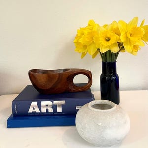 May include: A still life arrangement featuring a wooden bowl with a handle, a stack of blue books with the word "ART" in silver, a small white stone vase, and a vase of yellow daffodils in a dark blue vase.