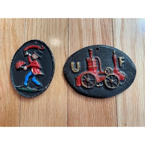 May include: Two cast iron wall plaques. One is oval-shaped with a figure in a red coat, blue pants, and a red hat blowing a horn. The other is an oval with a red steam engine and the letters "U F".