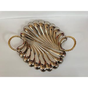 May include: A silver-toned metal serving tray shaped like a large scallop shell with a reflective, fluted surface. The tray features two circular, gold-toned twisted rope handles attached to each side, providing a contrast to the silver body.