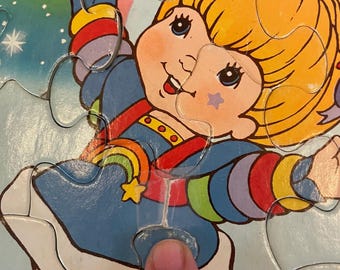 Puede incluir: Un colorido rompecabezas infantil con un personaje de dibujos animados con cabello amarillo y un vestido azul. El personaje está rodeado de un arcoíris y estrellas. El rompecabezas está hecho de cartón y tiene varias piezas.