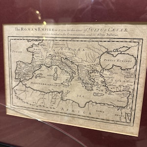 Op de afbeelding: Een ingelijste antieke kaart van het Romeinse Rijk ten tijde van Julius Caesar. De kaart, met gedetailleerde geografische kenmerken, bevat tekst bovenaan. De kaart is in zwart-wit, met tekst in een schreefletter.