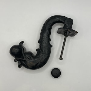 Puede incluir: Aldaba de puerta de hierro fundido negro con forma de criatura marina, posiblemente un pulpo o un calamar. La aldaba tiene un cuerpo curvado con detalles texturizados y una placa de montaje con un perno roscado. Una pequeña pieza redonda y negra está cerca.