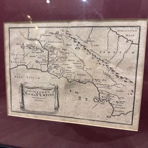 Può includere: Stampa di mappa antica dell'Impero Romano, incorniciata in un bordo rosso scuro. La mappa è dettagliata con caratteristiche geografiche, nomi di luoghi e testo, tra cui "THE INFANCY ROMAN EMPIRE according to Florus". La mappa è in bianco e nero.