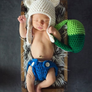 May include: A newborn baby wearing a white crocheted hat with bear ears and a blue crocheted diaper, lying in a wooden box lined with faux fur. The baby is holding a green and white crocheted toy.