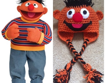 May include: An orange crochet hat with black hair, white eyes, a red nose, and a smiling mouth. The hat has ear flaps with black and orange braided ties ending in tassels. The image also shows a Sesame Street character, Ernie, wearing a striped sweater and blue jeans.