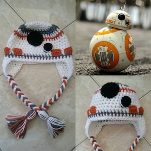 May include: A white, gray, and orange crocheted hat with black felt circles. The hat has a long, braided cord with tassels at the end. The hat is designed to look like the Star Wars character BB-8.